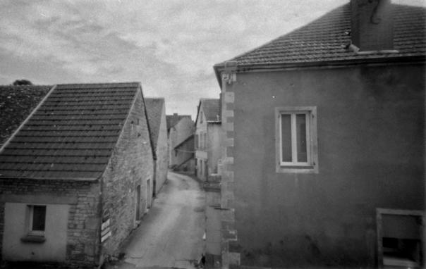 Photo noir et blanc depuis ma fenêtre. On vois la ruelle se perdre en face et des maisons, à droit et à gauche. Ciel nuageux d'un après midi à la fin mai.