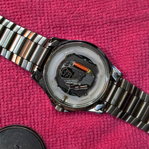 The opened back of a stainless steel Casio Quartz watch showing the movement and the battery. Pink cloth as background.