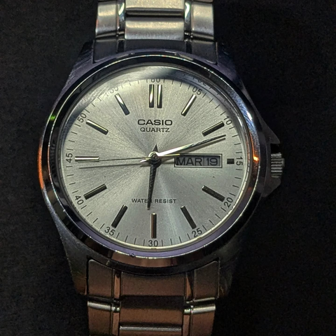 Frontview of a stainless steel Casio Quartz analog watch with a calendar complication. Has a brushed metal dial and hands.