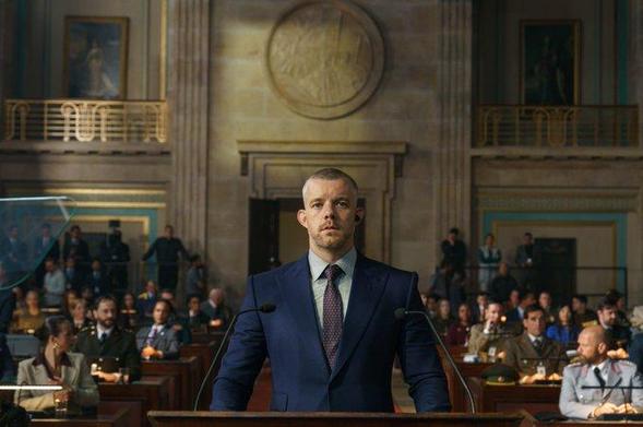 Russell Tovey as his character Barclay in a courtroom full of people. He wears a dark blue suit, light blue shirt and dark tie. He seems very stern and serious.