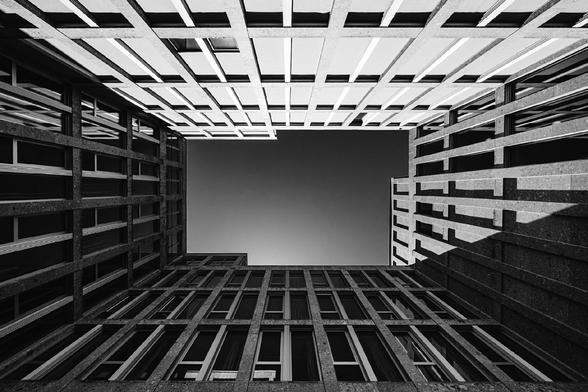 Looking up from a courtyard, the symmetrical walls of a modern building frame the sky in sharp geometric lines. The black and white contrast emphasizes the repeating windows and grid-like patterns of the concrete structure.

