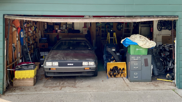 Photograph of the front of a DeLorean sports car pointed out of a crowded garage with bins, a snowblower, filing cabinets, tools, and other garage clutter visible.