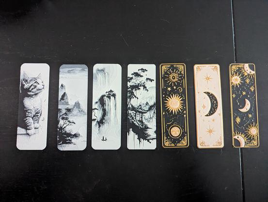 Seven 3d printed bookmarks. They're all basic black with colour over top. One is a black & white kitten, three are Japan inspired scenes (also black and white) and three are black/white/gold with a sun and stars motif. 