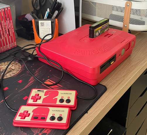 On peut voir au centre de l'image une Une Twin Famicom rouge avec le jeu Xevious chargé. Les deux manettes se trouvent sur le base de l'image. La console est posé sur un bureau.