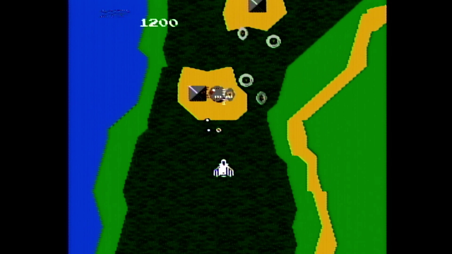 Capture d'écran d'une partie de Xevious sur Twin Famicom. Le décors est princiaplement vert avec un petit vaisseau blanc qui tire sur des ennemis en gris qui viennent du haut de l'écran