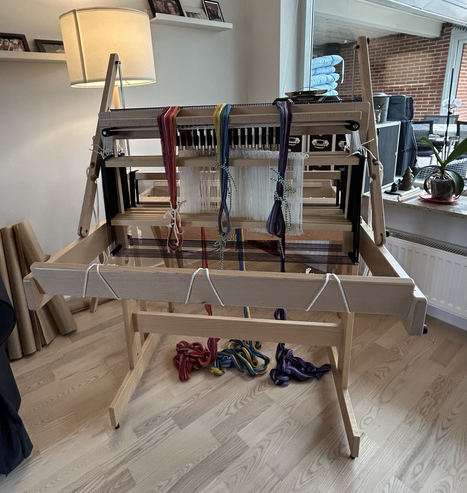 A look at the back side of a Louet Jane handloom with three warp chains in a rainbow of colours laid across the top of the loom 