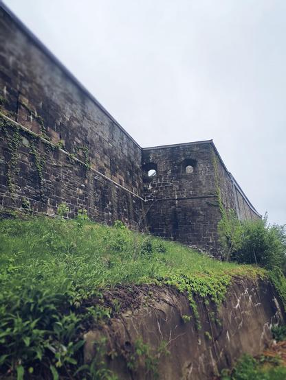 Vista lateral de la muralla de norte de la ciudad. Esta es de piedra, tiene dos ventanitas en una de sus caras y su base está llena de maleza fresca por la lluvia.