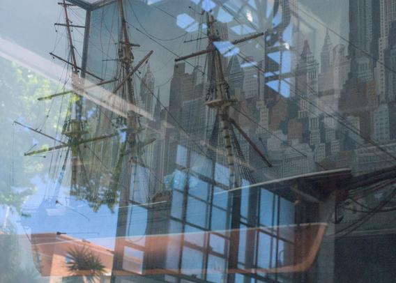 A diorama of a sailing ship model in front of a cityscape. There are strong reflections of factory windows on the glass case 