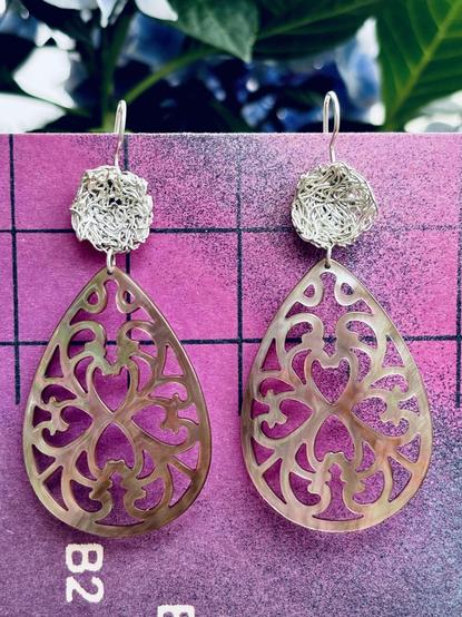A pair of ornate earrings featuring a teardrop shape with intricate cut-out designs resembling hearts and floral patterns. The top of each earring has a textured round component. The background is a vibrant pink with grid lines.