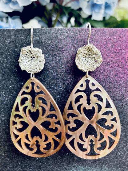 A pair of intricately designed drop earrings featuring a filigree pattern in shades of rose and brown, with a woven circular accent at the top. The earrings hang against a textured dark backdrop, complemented by soft, blurred floral elements in the background.