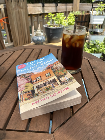 A class of iced pour over sitting next to a novel (welcome to the hyunam-dong bookshop by heart Bo-reum) on a table outside