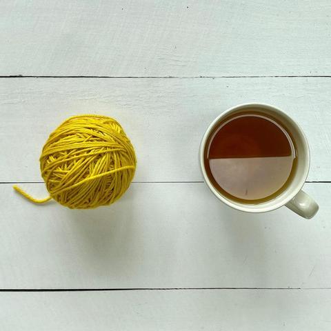Flat lay view of a bright yellow ball of yarn next to a large cup of tea on a white plank background.