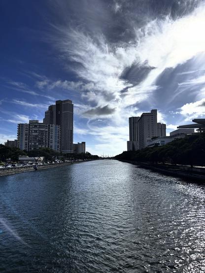 A serene canal reflecting a blue sky with wispy clouds. Tall buildings line the edges, and a tree-lined walkway runs alongside the water. The scene conveys a peaceful urban environment under bright daylight.