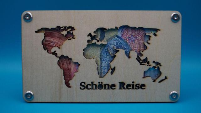 Geldgeschenkrahmen aus Holz mit gelaserter Weltkarte und der Aufschrift „Schöne Reise“, hinterlegt mit einem Geldschein.

