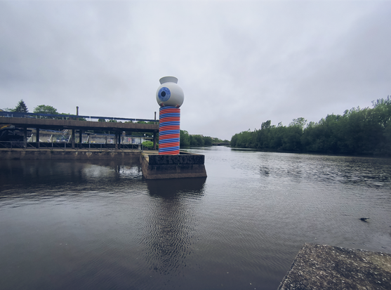 Vista al nivel del agua de la rivière St-Charles. En el centro de la imagen hay una escultura de un ojo sobre un cilindro metálico rayado rojo y azul.