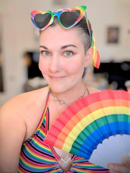 Cute woman wearing rainbow sunglasses, a rainbow hair tie, a rainbow crop top with a metal heart in the center while holding a rainbow fan.