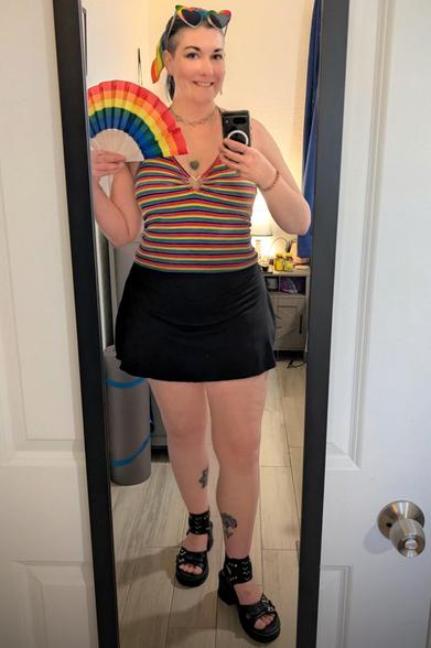 Cute woman wearing rainbow sunglasses, a rainbow hair tie, a rainbow crop top with a metal heart in the center while holding a