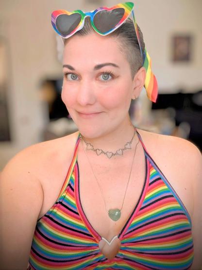 Cute woman wearing rainbow sunglasses, a rainbow hair tie, a rainbow crop top with a metal heart in the center.