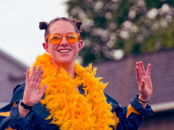 A woman wearing yellow glasses has her hair tied up up in two small buns, is smiling and is wearing a blue shirt with yellow stars and wearing something like a yellow lei and holds her hands up.