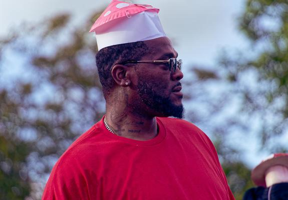A black man looks to the left with a funny white and red mushroom cap on his hed and a red shirt with blurred out leaves and branches behind him.