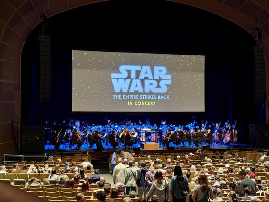 A concert hall is set up for "Star Wars: The Empire Strikes Back in Concert." The stage features an orchestra and a large screen displaying the title of the movie. The seating area is filled with an audience eagerly awaiting the performance.