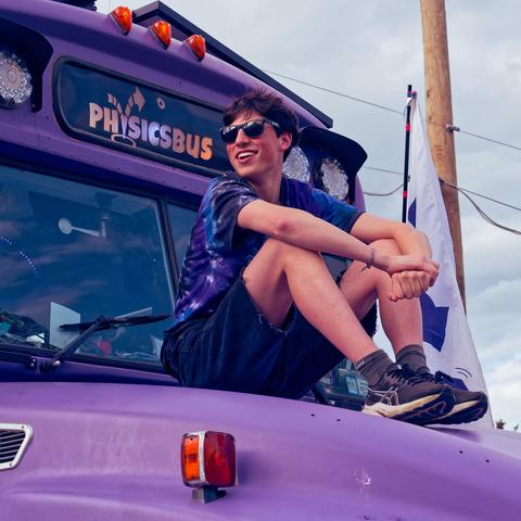 Teen boy with blue clothes and dark glasses rides on hood of a purple school bus where the route sign say PHYSICSBUS