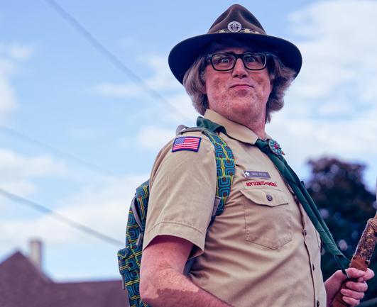 Man in boy scouts uniform wearing a us flag patch on his sleeves and a blue and green patterned backpack and glasses and a comical facial expression with grey hair and a fedora like hat