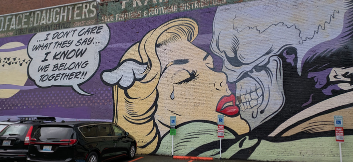 A classic comic-style mural, about 20 meters wide and 6 meters high, shows a skull-faced individual and a blonde, lipstick-wearing woman, tears dripping from her closes eyes, embracing, maybe kissing. She says, "I won't care what they say... I know we belong together!"