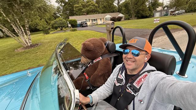 A person in a blue roadster car, taking a picture when driving, with a teddy bear in the passenger seat