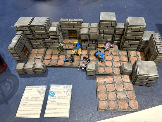 A playfield for a miniatures skirmish game. The photo shows a dungeon room with gray stone walls and brown floor tiles. Several miniature figures lie fallen on the floor - two cultists in blue robes, two heros, and one zombie. The two remaining figures are squaring off, an archer and a cultist advancing on him.