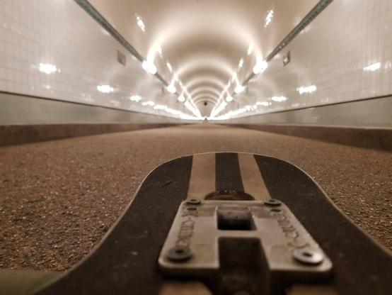 Bildbeschreibung von KI:
Das Foto zeigt eine ungewöhnliche Perspektive aus dem Alten Elbtunnel in Hamburg – aufgenommen in Bodennähe, direkt hinter einem Longboard oder Skateboard, das im Vordergrund liegt. Der Blick geht in die Tiefe des Tunnels.

Die Wände des Tunnels sind mit weißen Kacheln bedeckt, die durch die symmetrisch angebrachten, runden Wandlampen gleichmäßig beleuchtet werden. Die Beleuchtung erzeugt eine rhythmische Abfolge aus Lichtreflexionen, die sich entlang der gekachelten Tunnelwände nach hinten verjüngen – was dem Bild eine starke Tiefenwirkung verleiht. Der Tunnel ist leicht gebogen und wirkt dadurch fast endlos.

Der Boden ist asphaltiert oder rau asphaltiert – typisch für eine Fußgänger- oder Fahrradverbindung. Das Longboard im Vordergrund wirkt benutzt, das Metallteil ist leicht angerostet. Die Kamera ist so niedrig positioniert, dass man aus Sicht des Boards quasi nach vorn schaut – als würde es gleich durch den Tunnel rollen.

Das Bild vermittelt ein Gefühl von Weite, Perspektive und Urbanität – mit einer Prise Abenteuer. 