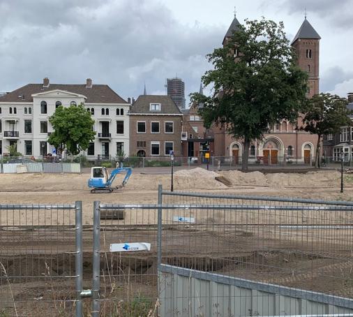 Sand field with small excavator, construction fencing. In the background facades of buildings, a tree.