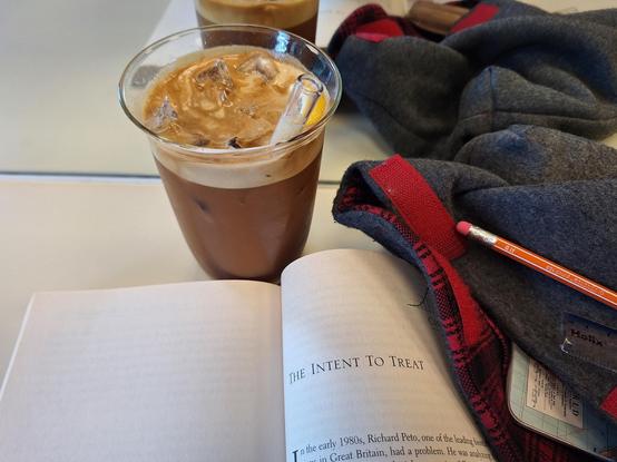 Glass with a brownish fluid and a glass straw. Ice cubes floating on top. The glass stands in front of a mirror on a counter or table top, next to a book* opened on a page with the heading "The intent to treat". Other stationary is visible and som grey, red, black tartan cloth.

*David Salsburg, The lady tasting tea