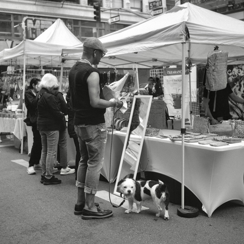 A black and white film photo. A man, with his dog, is looking over a bag at the farmers market in downtown Los Angeles, California. Sunday, May 18, 2025.