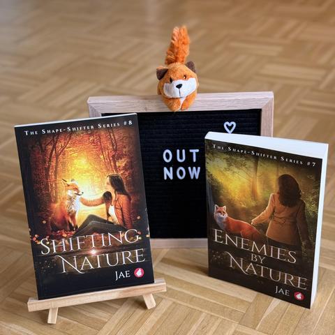 Shown are paperback copies of Enemies by Nature and Shifting Nature by Jae. They are flanking a board that says "out now." On top of the board sits a tiny stuffed fox. The covers depict a brunette woman and a red fox facing each other in the forest. 