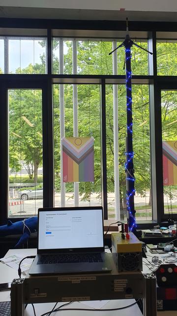 Tisch voller Technik, in der Mitte ein blau beleuchteter Antennenmast und im Vordergrund ein selbstbauter Sender im Rack, darauf ein Laptop mit Control Panel bzw. Eingabemaske für zu sendene Nachrichten