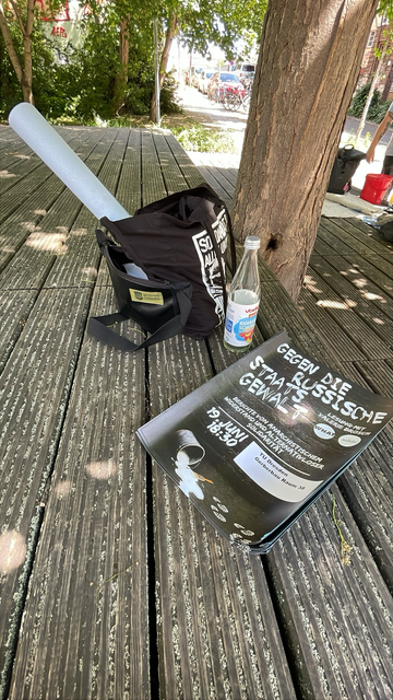 Auf einer Bank liegt ein kleiner Stapel Plakate. Auf dem Plakat steht „Gegen die russische Staatsgewalt. Berichte von anarchistischem Widerstand und alternativloser Solidarität. Lesung mit Valerie Brosch. WHAT, Solida. 19. Juni 18:30 Uhr, TU Dresden Gerberbau Raum 38.“