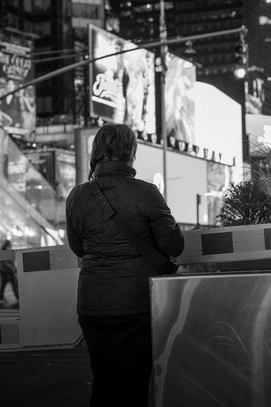 Black and white photo of a person standing with their back to the camera in Times Square, New York City. The person wears a quilted jacket and is framed against bright lights, signage, and city structures, creating a contrast between motion and stillness.