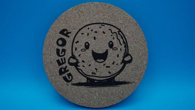 Runder Korkuntersetzer mit einem lachenden Donut-Motiv und dem Namen „Gregor“, vor blauem Hintergrund leicht schräg fotografiert.