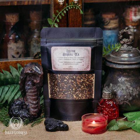 A black pouch of dried herbs sits on a burlap-covered surface. There is a window in the front and a label that reads, "Lilith Herbal Tea" with a list of ingredients: black tea, cinnamon, ginger, cardamom, clove, and pepper. This is an exclusive product from Inked Goddess Creations.