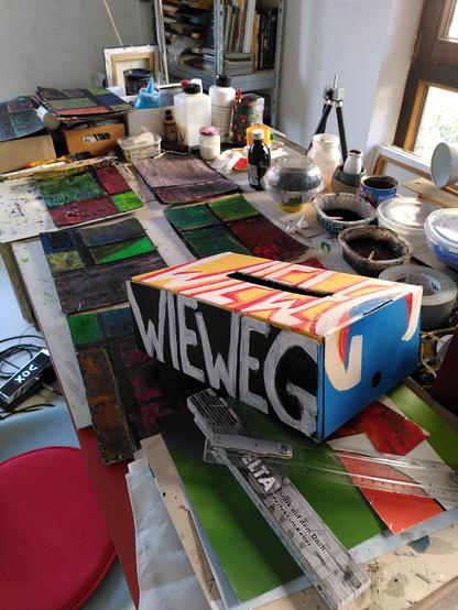 Ein Künstler-Arbeitsplatz, darauf eine bunt bemalte Spendenbox, auf der in dickem Pinselstrich „Wieweg“ steht.