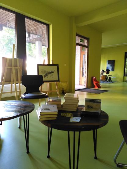 Ein limettenfarbener, großer Raum, in dem ein Büchertisch mit Fees Werken steht. Außerdem Staffeleien und ein Schlagzeug.