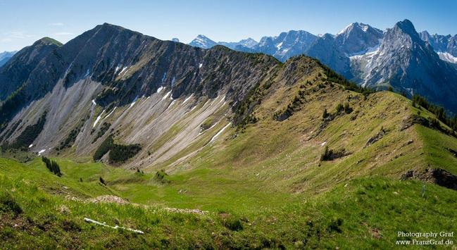 A serene landscape featuring a green mountain with snow on the side, set against a backdrop of a clear blue sky. The mountain range is rugged and rocky, with patches of green grass and trees scattered throughout. The scene exudes a sense of tranquility and majesty, showcasing the beauty of nature in its raw form. The snow-capped peaks contrast beautifully with the greenery below, creating a stunning visual contrast. This image captures the essence of outdoor adventure and the peacefulness of un…