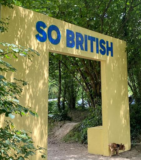 In a wooded setting a yellow square arch is emblazoned atop with the words "SO BRITISH" in blue.  At the bottom right a small tricolour terrier type dog is peeing on the arch 