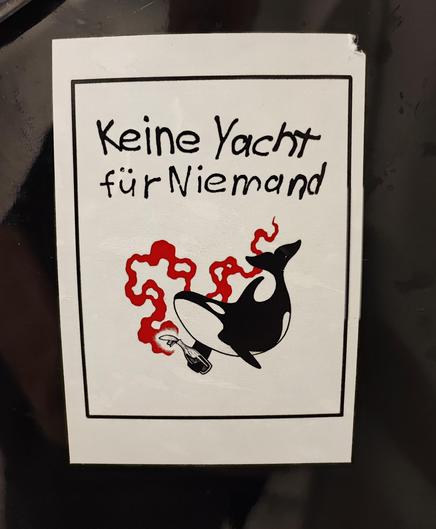 Foto eines Aufklebers auf schwarzem Hintergrund, weißer Aufkleber im Stil des Plattencovers von Ton Steine Scherben diesmal nicht mit dem Titel Keine Macht für Niemand, sondern Keine Yacht für Niemand und einem Orka mit Molotovkocktail in der Flosse.