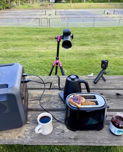 A behind the scenes shot of the set up for a YouTube video. In the foreground a portable power station, a toaster, an electric kettle, a jar of jam, some toast and a cup of coffee. Everything sitting on an old, wooden picnic table. In the middle of the frame is a camera pointed towards the all the elements on the table. A second, action camera in on the right hand side. The background is a grassy field with a tennis court at the top end of the frame. The set up was to record a video on how a portable power station can elevate your camping game.