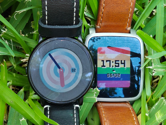 Two Pebble Time smartwatches side by side on a grassy backdrop. The round watch to the left shows an analog clock face with concentric rings in transgender pride colors, and the rectangular watch to the right shows a digital clock face backed by a six-stripe rainbow flag.