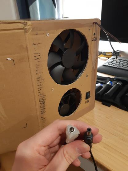A DIY solder fume extractor project in progress. The fume extractor is made from a box that has two holes with fans in them. Next to the upper fan is a button titled "LED" and two exposed wires (intended for an LED strip). Next to the lower fan is a flip switch titled "FAN" that is set to off.
In focus in the picture are a male and a female barrel jack connector end with the female one being completely DIYed and held together with hot glue.
