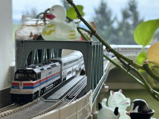 An Amtrak EMD SDP40F model locomotive, a boxy diesel locomotive, mostly silver with some black elements and a red stripe and blue stripe running along its entire length, pulling four silver double-decker passenger cars, on a double-track model railroad running over a truss bridge. A pothos vine wraps over and next to the bridge. A few halloween-themed rubber ducks are placed on the green artificial turf on the desk next to the bridge, and one, white with green bat silhouettes, sits on top of the bridge. A window is in the background, with blurry trees visible in the distance.