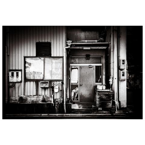A gritty exterior of what appears to be a back entrance or side façade of a building in Nagoya. The image is rendered in high-contrast black and white, drawing attention to the weathered siding, a rust-streaked window, and a dented metal door with a screen panel. To the right, stacks of wooden boards are topped with a couple of plastic buckets, hinting at some kind of industrious activity—past or present. Pipes, meters, and wires trace their way across the surfaces like urban veins, adding a quiet complexity to the otherwise utilitarian composition.